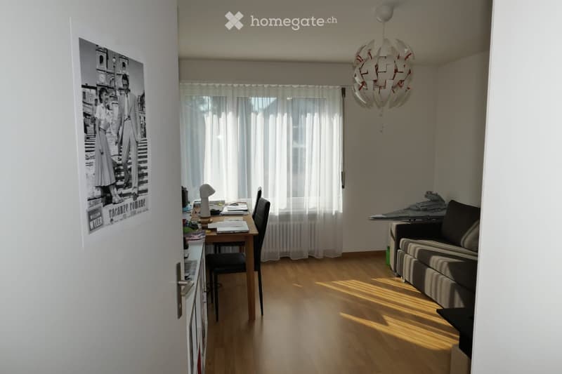 Bright, open living space with wooden flooring, large windows with white curtains, a modern chandelier, a sofa, a dining table with chairs, and a desk area with a lamp.