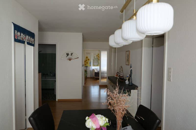 A modern hallway leading to a dining area with a black table, chairs, and decorative vases. The hallway features white walls, wooden flooring, and a hanging light fixture.