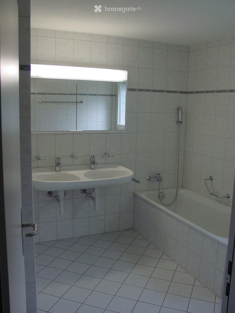 Clean bathroom with white tiled walls and floor, featuring a double sink, bathtub, and large mirror with built-in lighting.