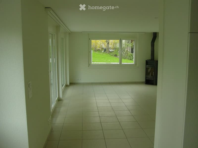 Empty room with tiled floor, white walls, large window, and a small stove in the corner.