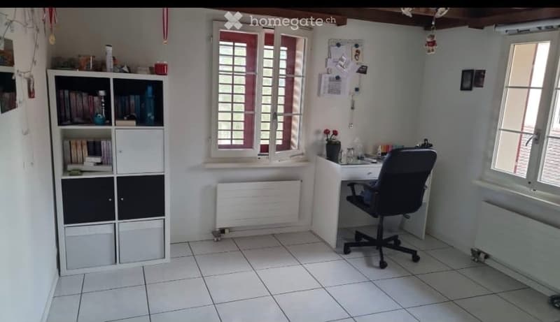 A tidy, well-lit room with white tiled flooring, a large window with white shutters, a bookshelf filled with books and decorative items, a desk with a chair, and a small flower pot on the windowsill.