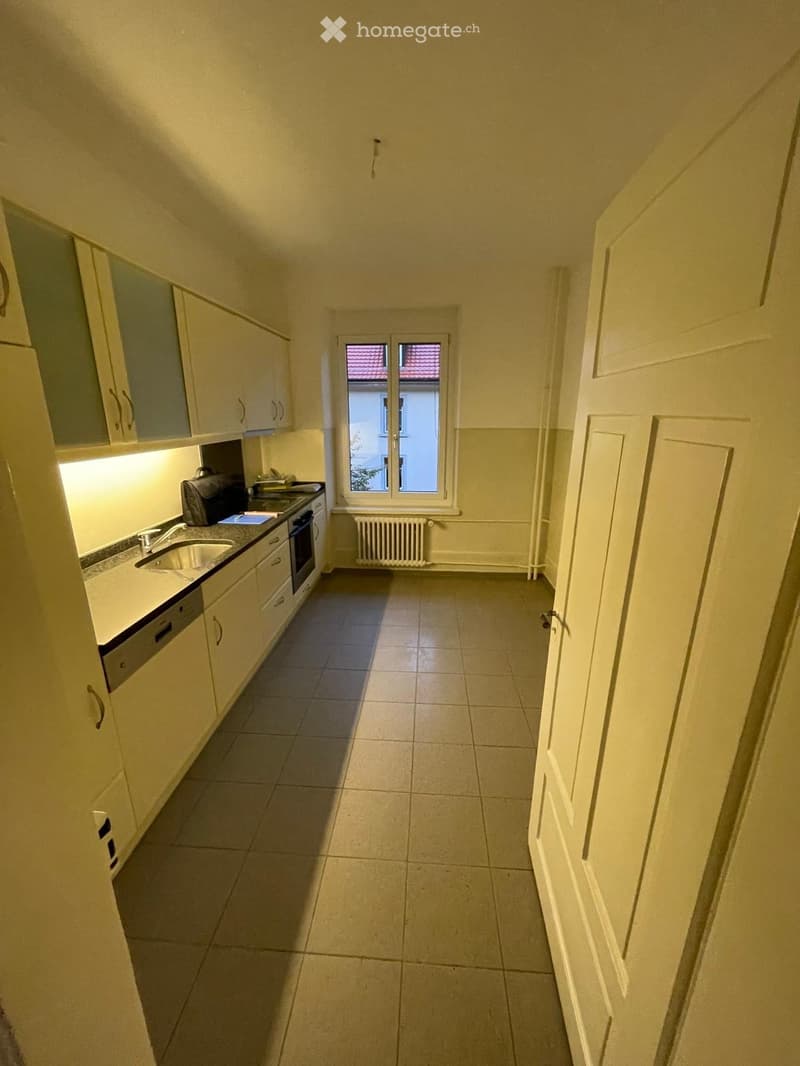 Modern kitchen with white cabinets, a sink, oven, and dishwasher. The space is clean and well-lit with a tiled floor and a window showing a view of a neighboring building.