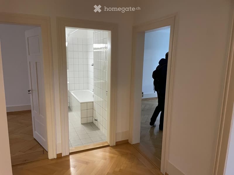 Hallway with wooden flooring, leading to a bathroom with a bathtub and tiled walls, and another room with a person partially visible.
