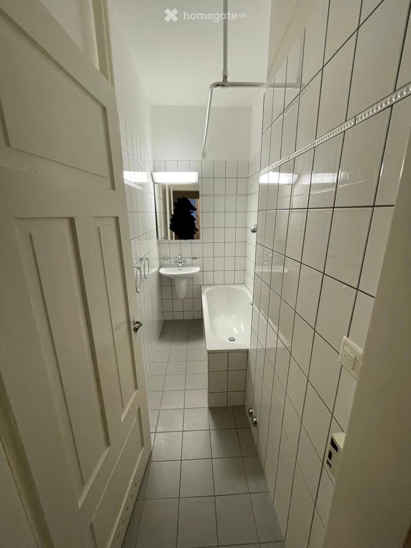 Small bathroom with white tiled walls and floor, featuring a bathtub, sink, and mirror.