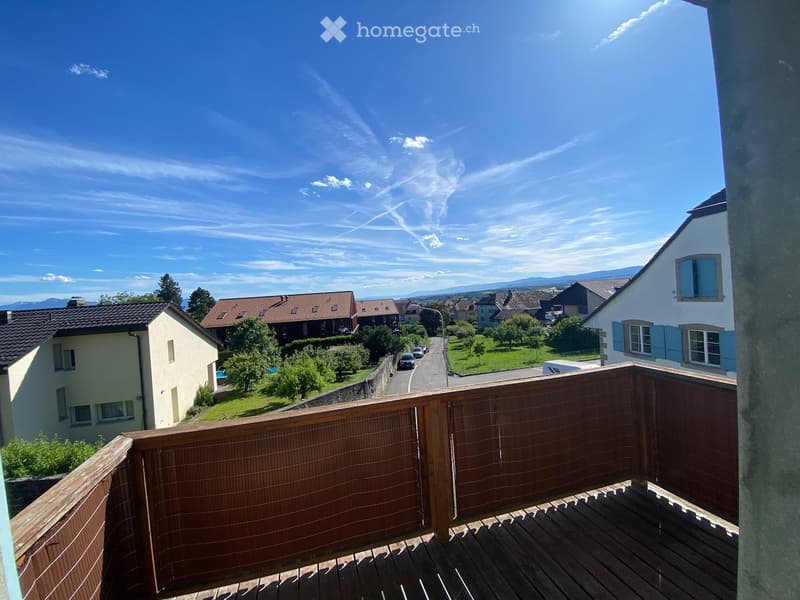 Balcony with wooden decking and railing, overlooking a residential neighborhood with houses, trees, and a clear blue sky.