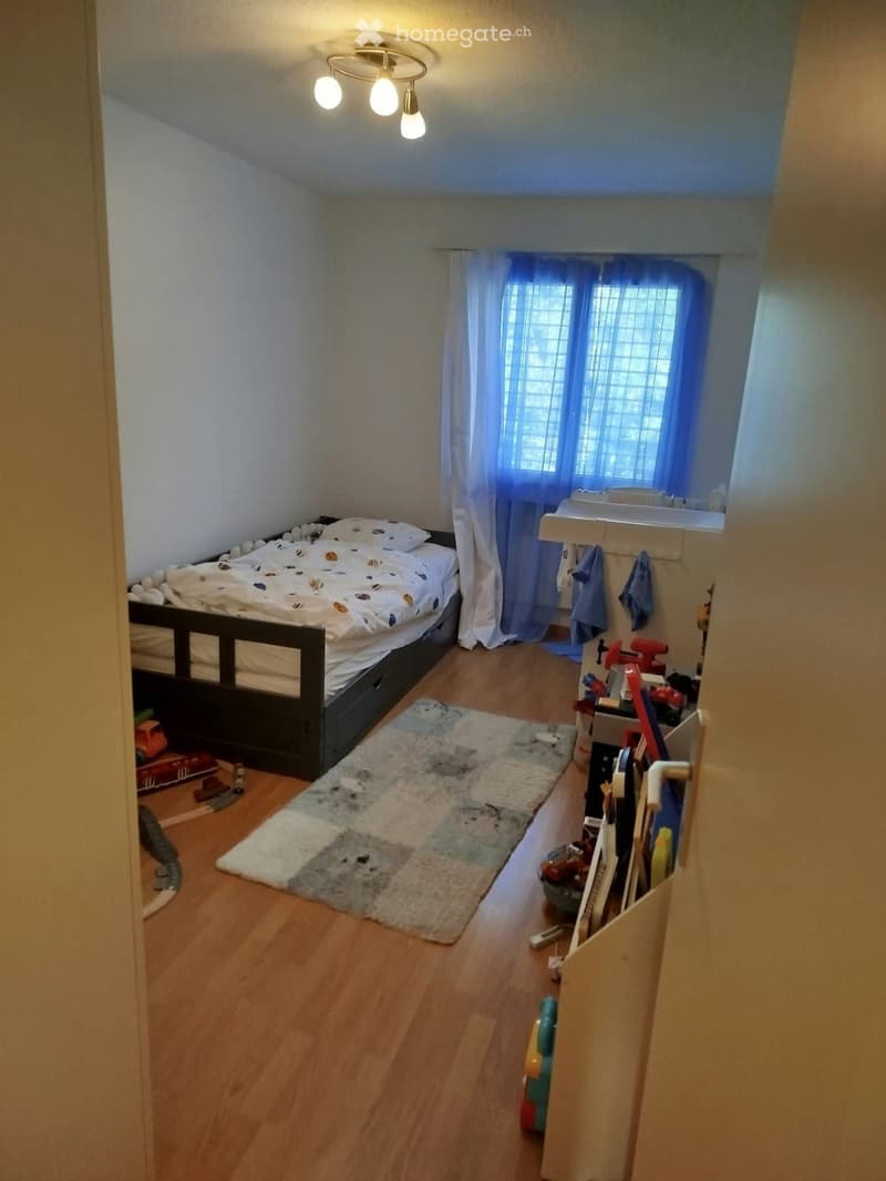 Cozy bedroom with a bed, patterned rug, small sink area, and toys visible. Wooden flooring and white walls.