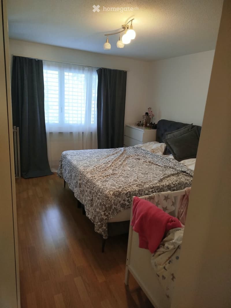 Cozy bedroom with wooden flooring, a double bed with patterned bedding, a nightstand, and a small baby crib with a red blanket.