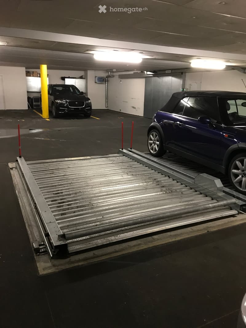 Underground parking area with two cars parked, one on a lift platform, and a yellow pillar and storage area visible.