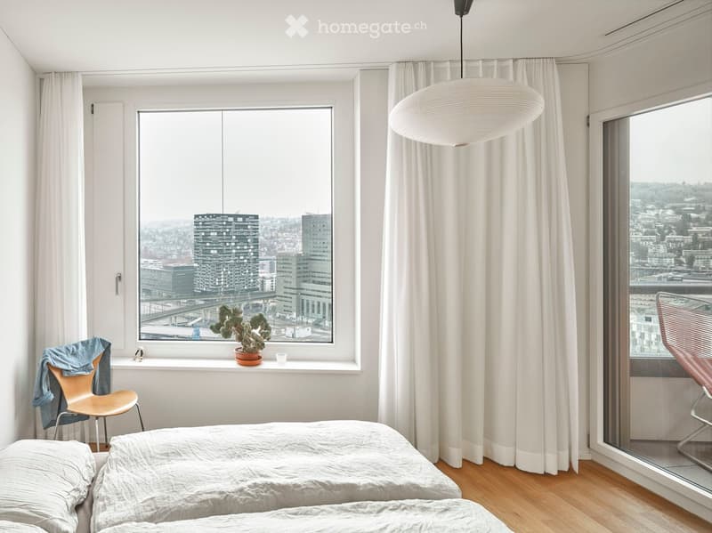Two single beds, wooden floor, white walls, large windows, white curtains, hanging lamp, potted plant on window sill, chair, blue jacket on chair, city view