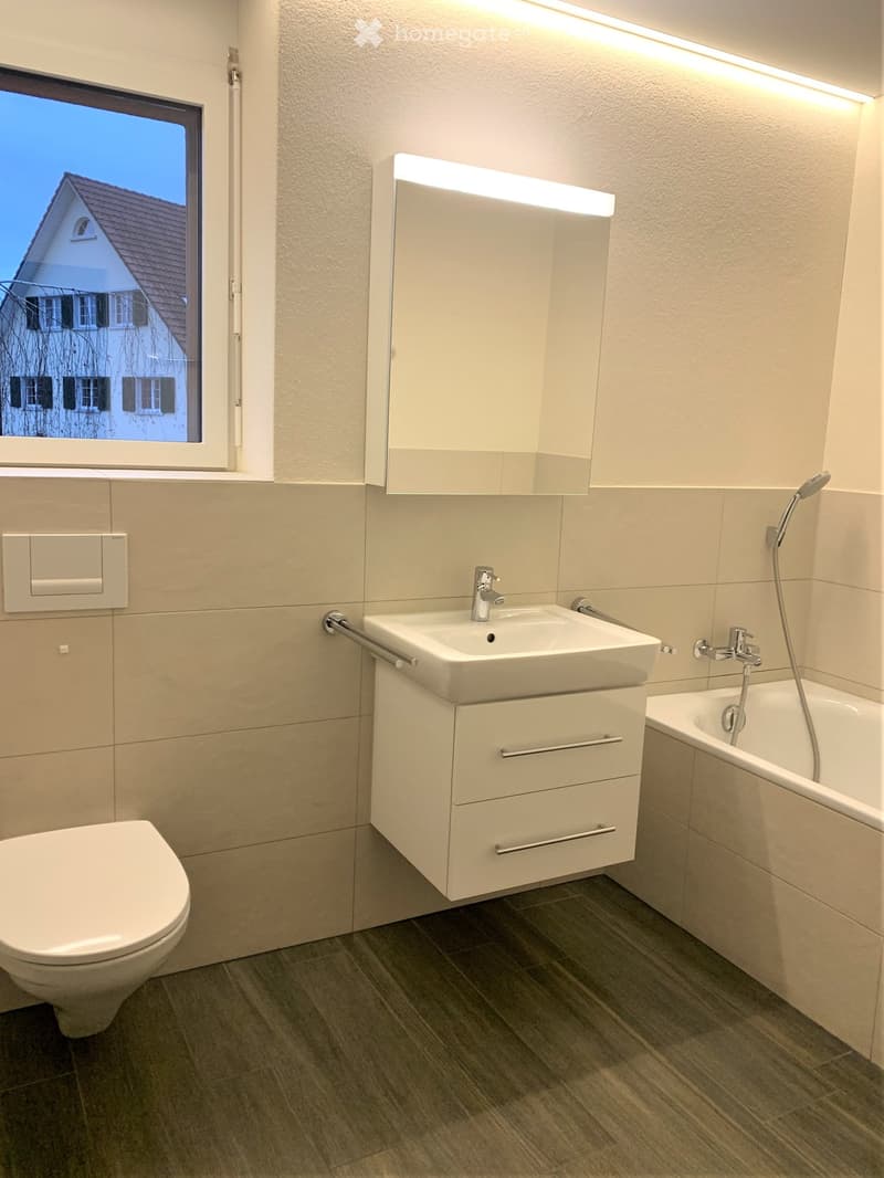 Modern bathroom with a white toilet, sink with a cabinet, bathtub, and a mirrored cabinet with built-in lighting. The room features light-colored tiles and a large window showing a house exterior.