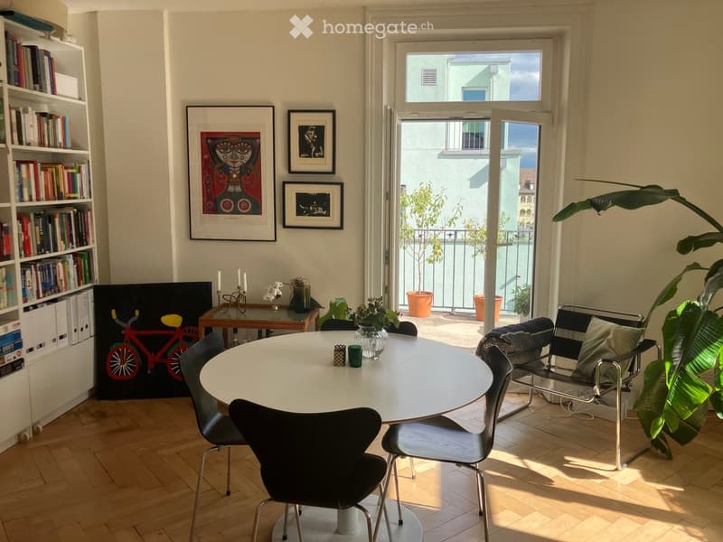 A modern, well-lit room with a round table surrounded by chairs, a bookshelf filled with books, a small table with a candle holder and vase, and large windows opening to a balcony with plants.
