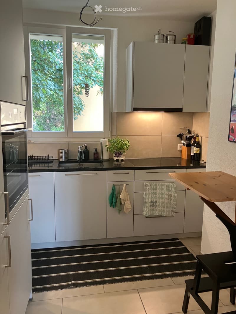 Modern kitchen with light grey cabinets, stainless steel appliances, a black countertop, and a small wooden table with a stool. Features a sink with a faucet, a towel rack with towels, and a potted plant on the counter.