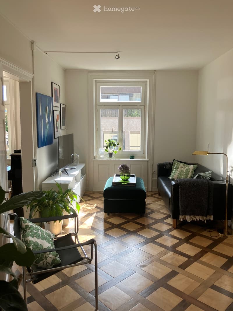 Living room with checkered tile flooring, large window with view, sofa, ottoman, armchair, coffee table, TV, and decorative plants.