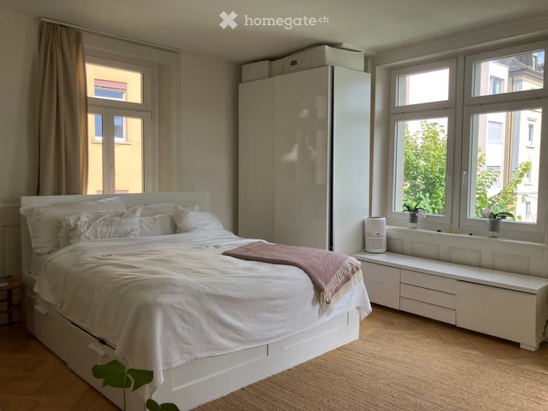 Bright bedroom with a white bed, nightstand, and large windows with curtains. Includes a radiator, fan, and potted plants on the windowsill.