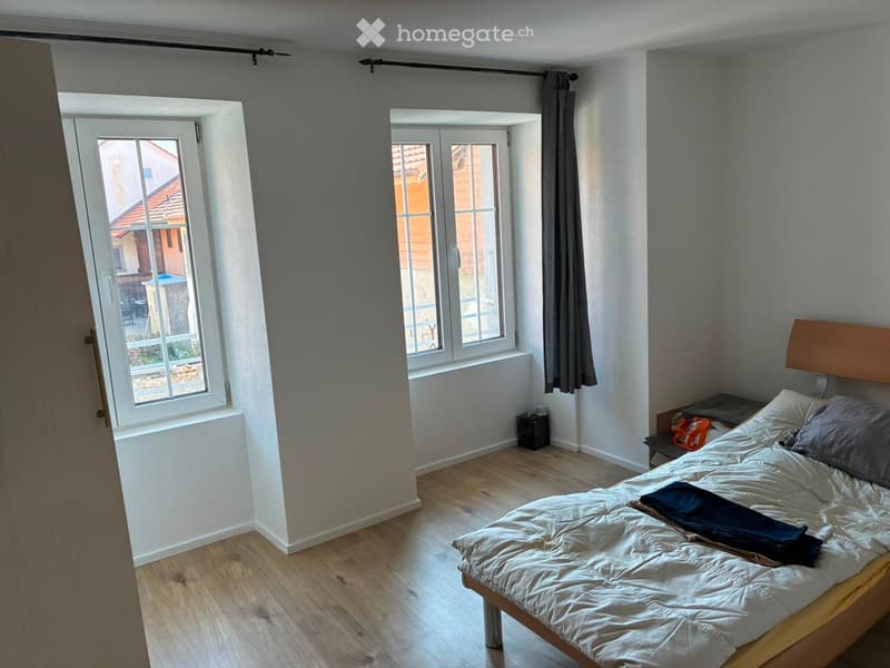 Bright bedroom with wooden flooring, large windows, a neatly made bed with pillows and a blanket, and a nightstand with a lamp.