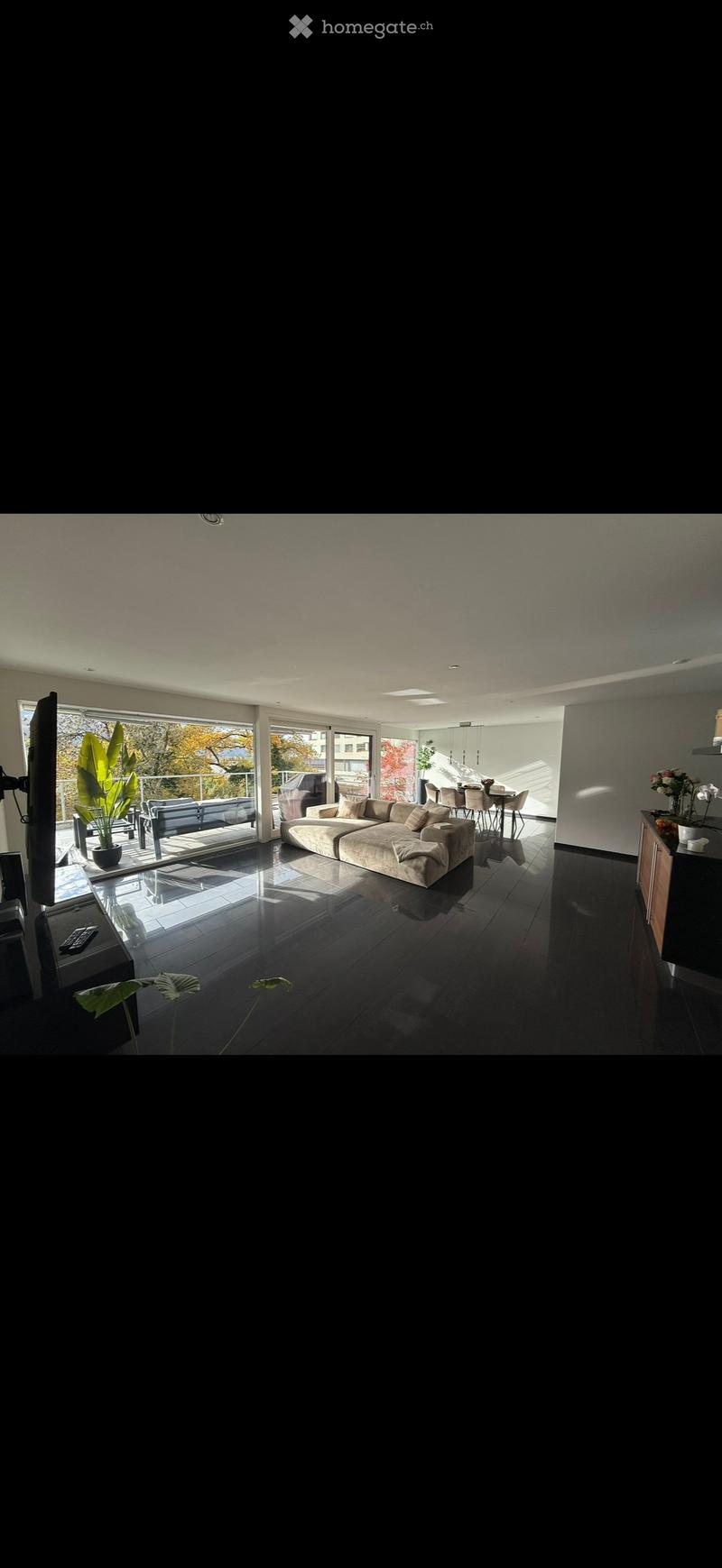Modern living room with large windows, dark tiled floor, beige sectional sofa, coffee table, dining area with chairs and table, television, and decorative plants.