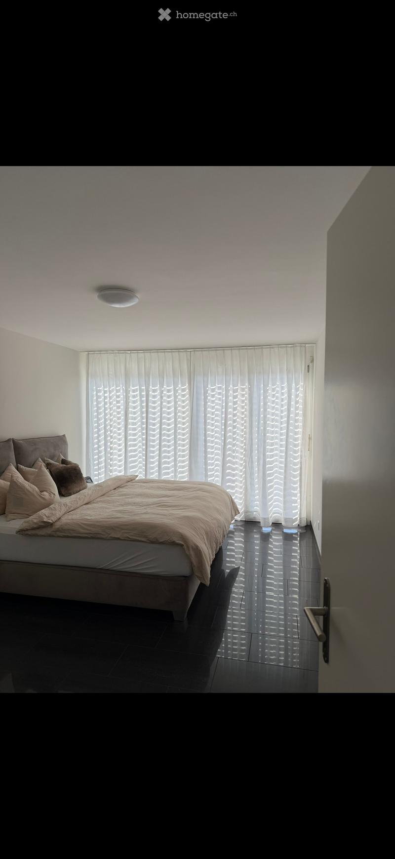 Modern bedroom with a large bed, multiple pillows, and light-colored bedding, set against dark tiled flooring and white walls with a large window covered by sheer curtains.