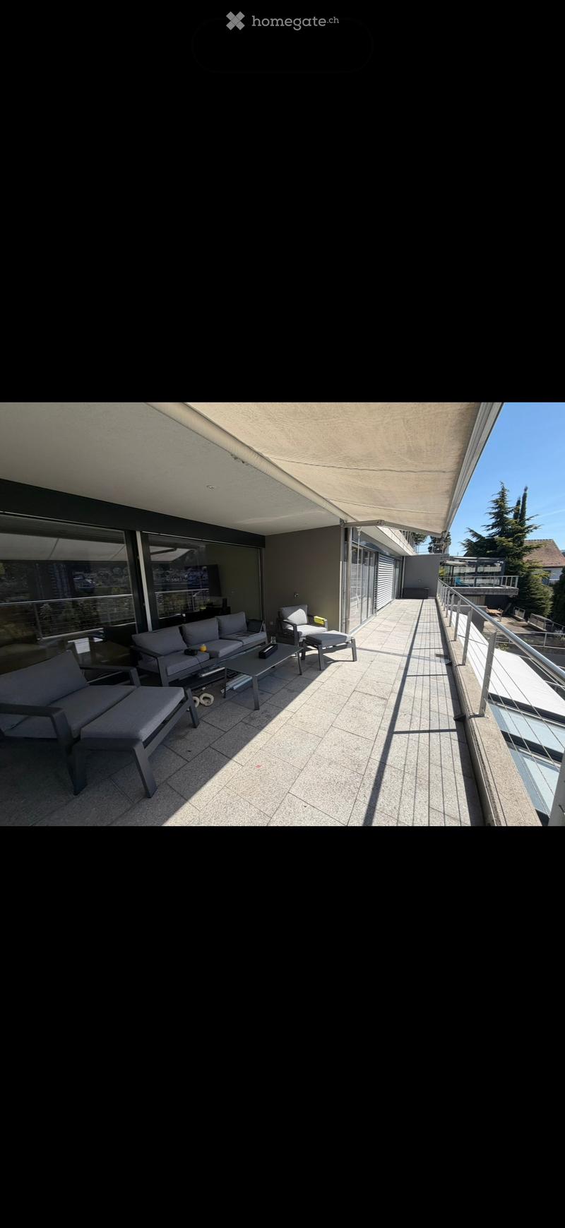 Modern balcony with grey sofa, coffee table, and chairs, featuring a retractable awning and a view of the surrounding area.