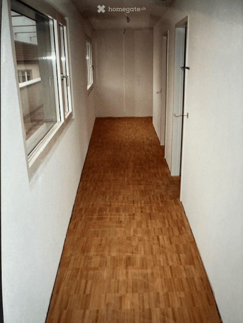Empty hallway with wooden flooring, white walls, and several doors and windows.