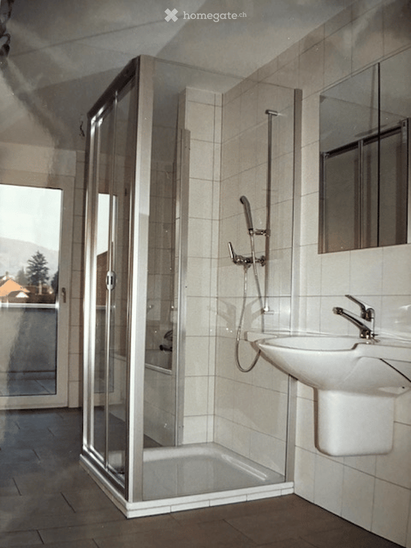 Modern bathroom with a glass-enclosed shower stall, a white sink with a chrome faucet, and a mirror above the sink. The walls are tiled in light beige tiles, and the floor is covered with brown tiles.