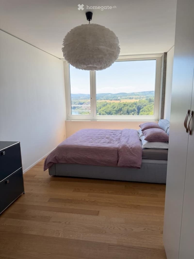 Modern bedroom with wooden flooring, large window offering a scenic view, a neatly made bed with pillows and a duvet, and a stylish pendant light fixture.