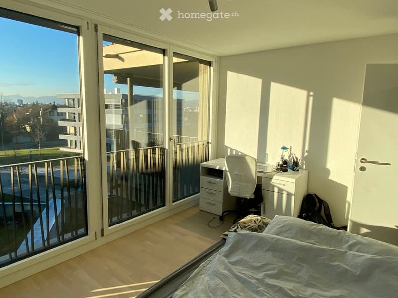 Bright bedroom with wooden flooring, large sliding glass doors leading to a balcony, a bed with pillows and a blanket, a desk with a chair, and a lamp on the desk.