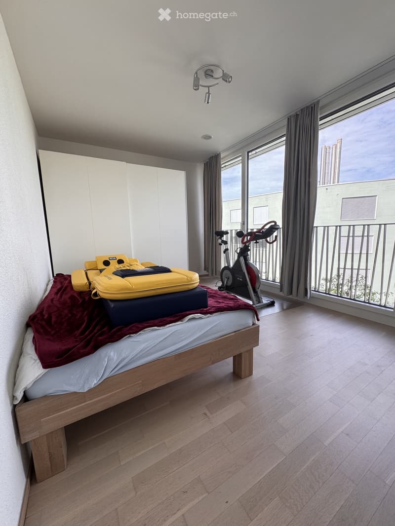 Modern bedroom with wooden flooring, large window with balcony access, exercise bike, and a neatly made bed with pillows and blankets.