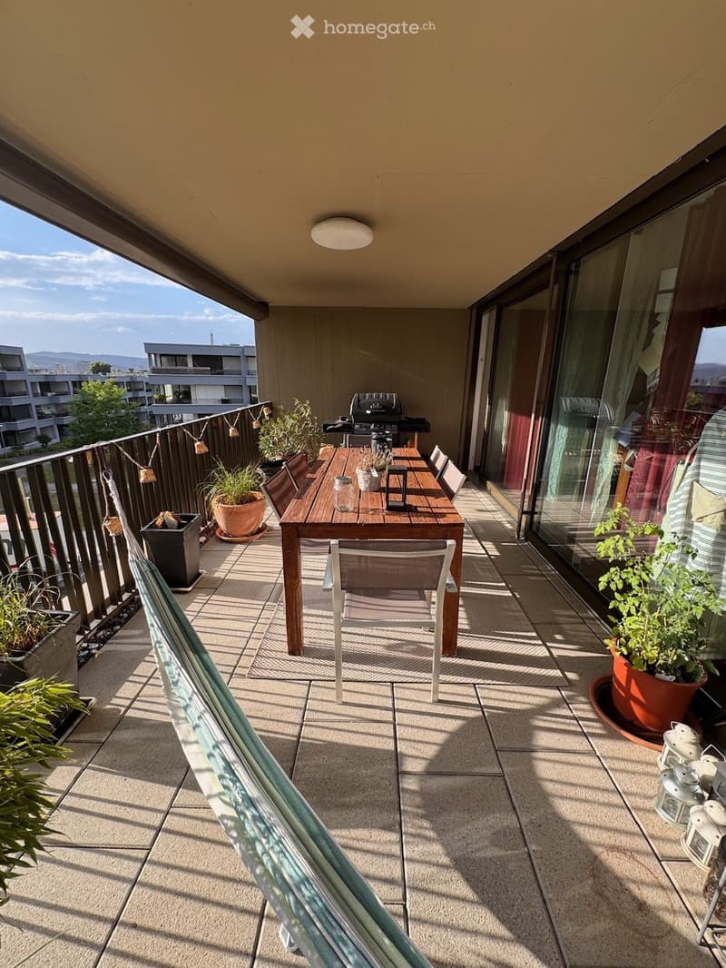 Balcony with a wooden dining table, chairs, potted plants, a hammock, and a grill, overlooking a cityscape.