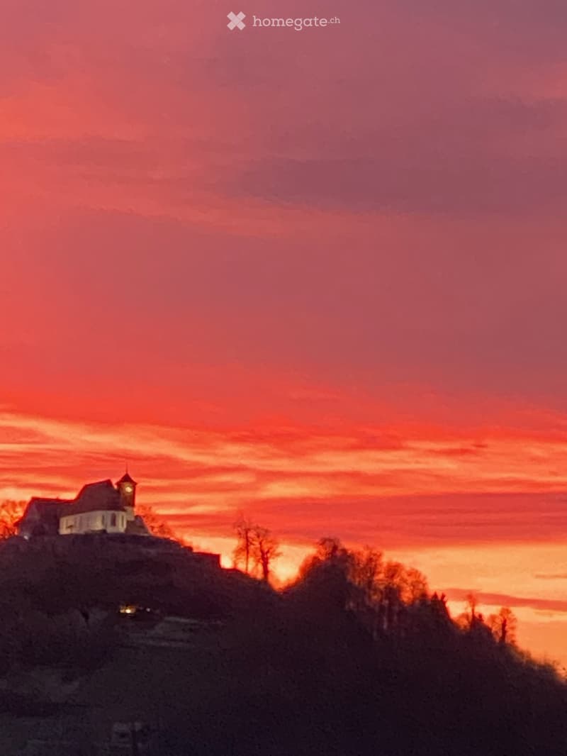 A silhouette of a building with a tower against a vibrant sunset sky, with trees and a distant body of water visible in the background.