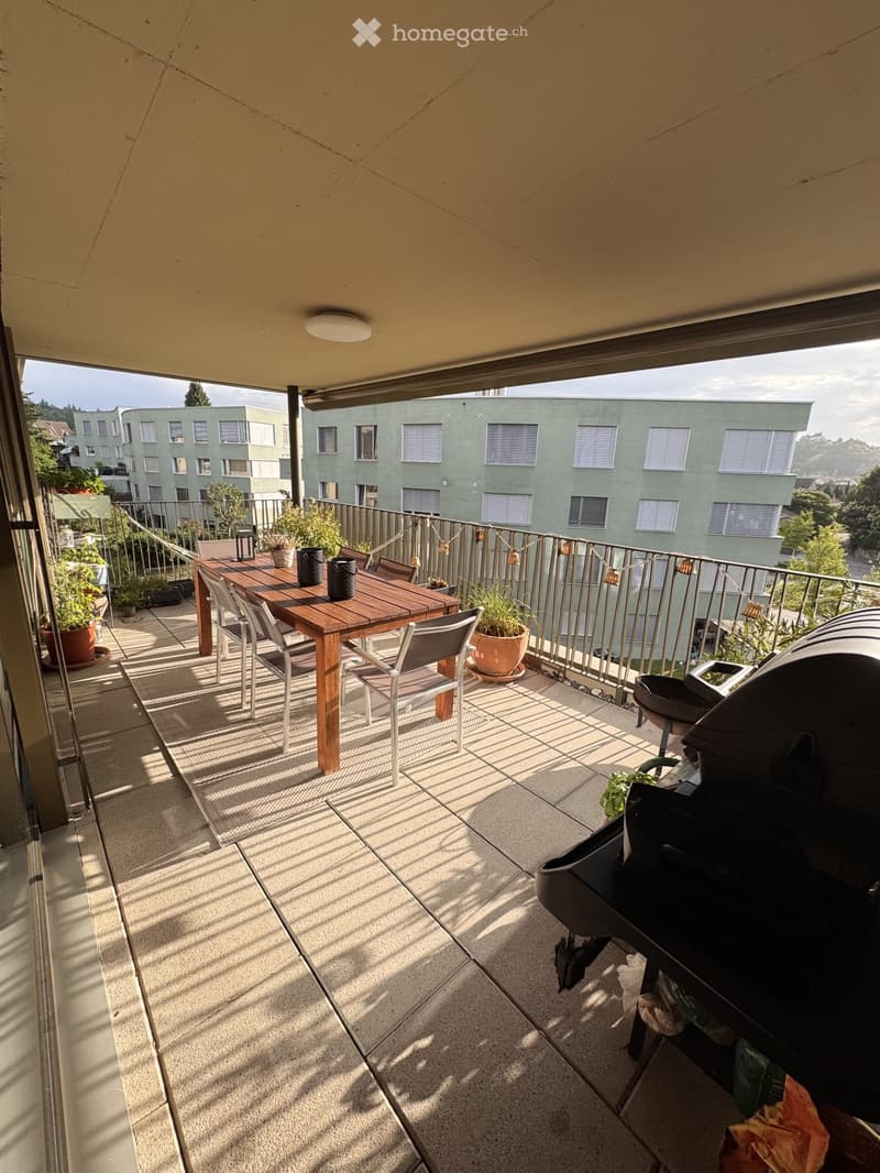 Balcony with a wooden table, chairs, potted plants, and a grill, overlooking a building with multiple windows.