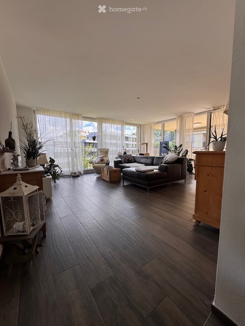 Modern living room with dark wooden flooring, large windows with sheer curtains, a black leather sofa with ottoman, armchair, side table, and decorative plants.