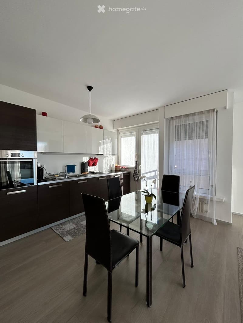 Modern kitchen with white and dark cabinets, stainless steel oven, sink, and a glass dining table with four black chairs. Natural light from large windows with white curtains.