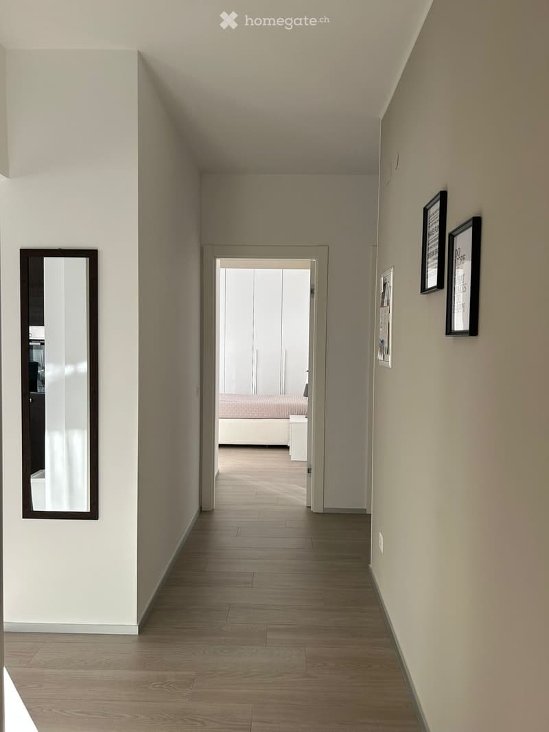 A narrow hallway with light wooden flooring, white walls, and a mirror on the left side. A door at the end leads to a bedroom with a visible bed and white furniture.