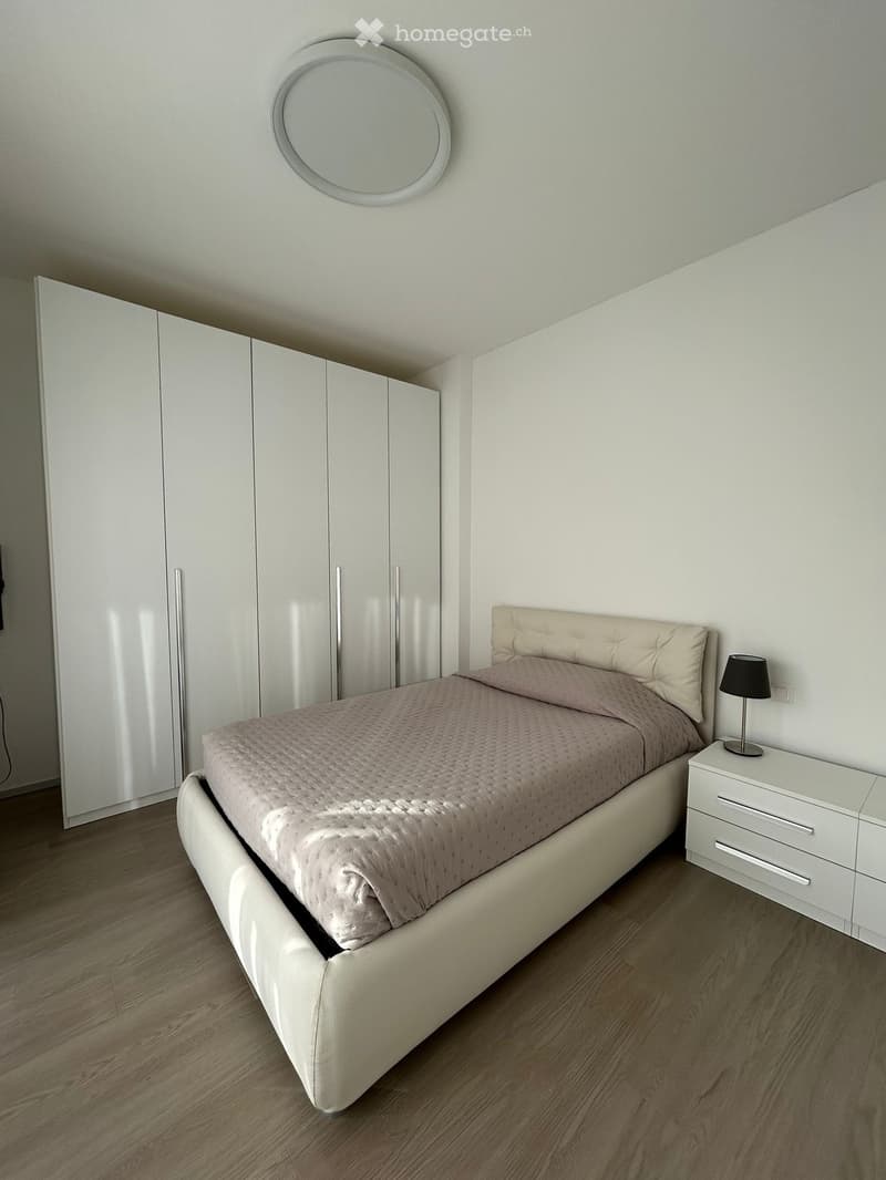 Modern bedroom with a white upholstered bed, beige bedding, white nightstand with a lamp, large white wardrobe, and a round wall-mounted mirror.