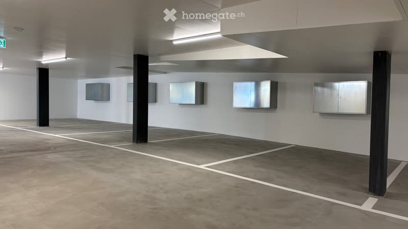 Empty underground parking area with concrete floors, white walls, and several metal cabinets mounted on the walls. The space is well-lit with fluorescent lighting and features structural pillars.