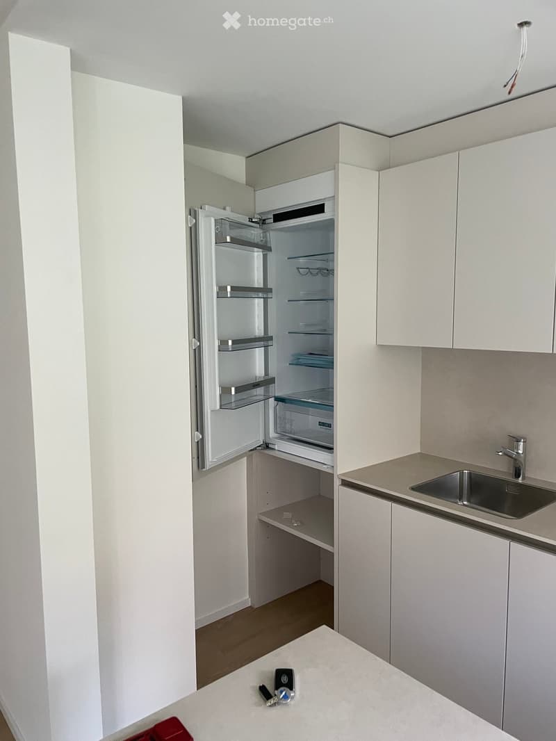 Modern kitchen with white cabinets, stainless steel sink, and an open refrigerator. Countertop has small items like keys and a remote control.