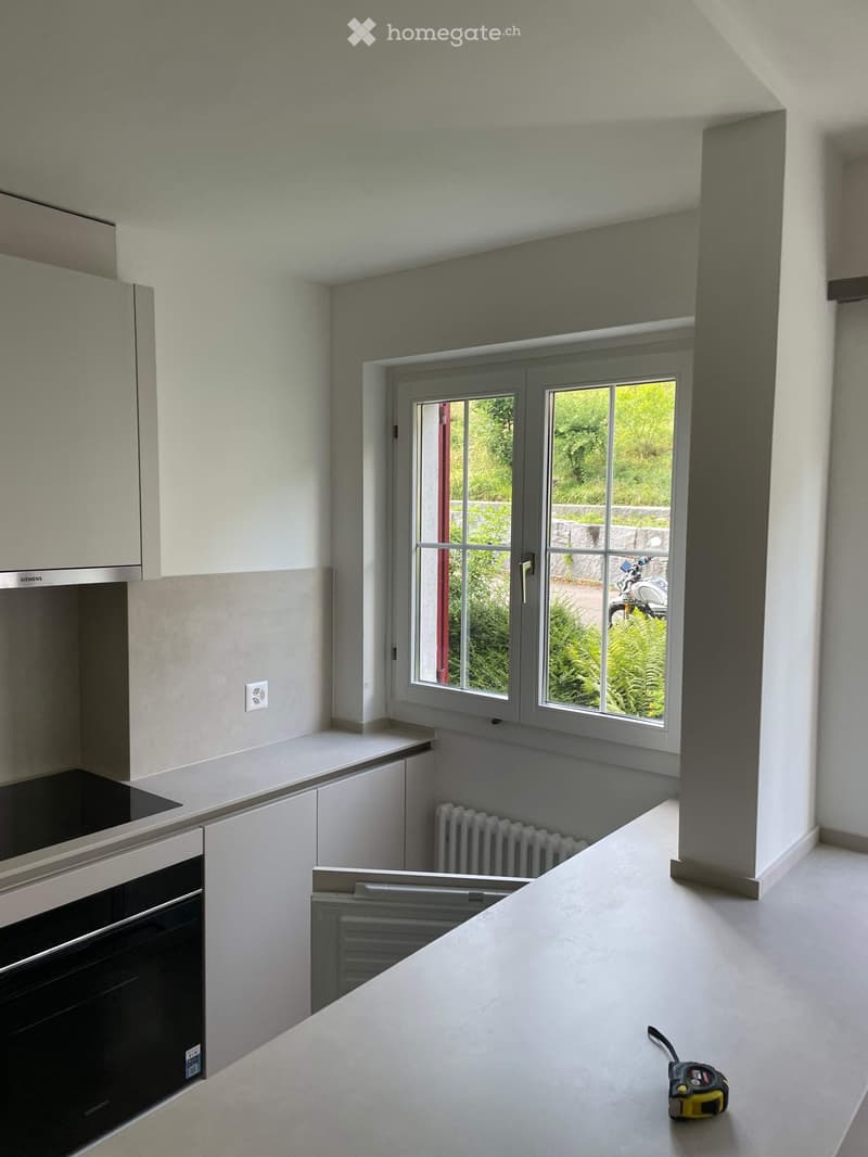 Modern kitchen with white cabinets, stainless steel oven, countertop, and a window overlooking a green area. A measuring tape is placed on the floor.