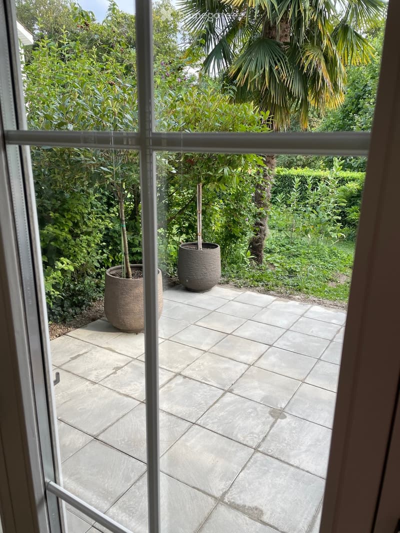 View from inside through a glass door onto a tiled balcony area surrounded by lush greenery and potted plants.
