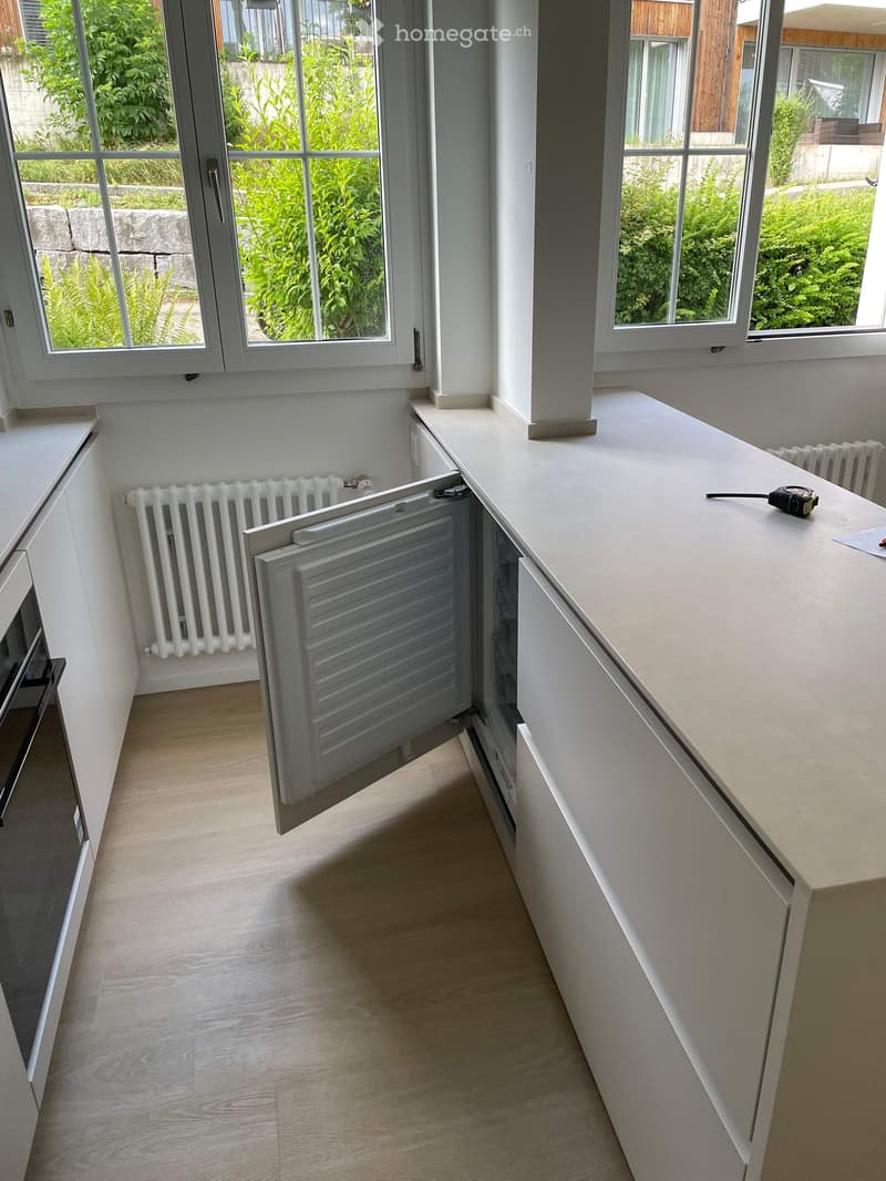 Modern kitchen with light-colored wooden flooring, white cabinets, and a long white countertop. A radiator is visible under the window, and an oven is partially visible on the left side.