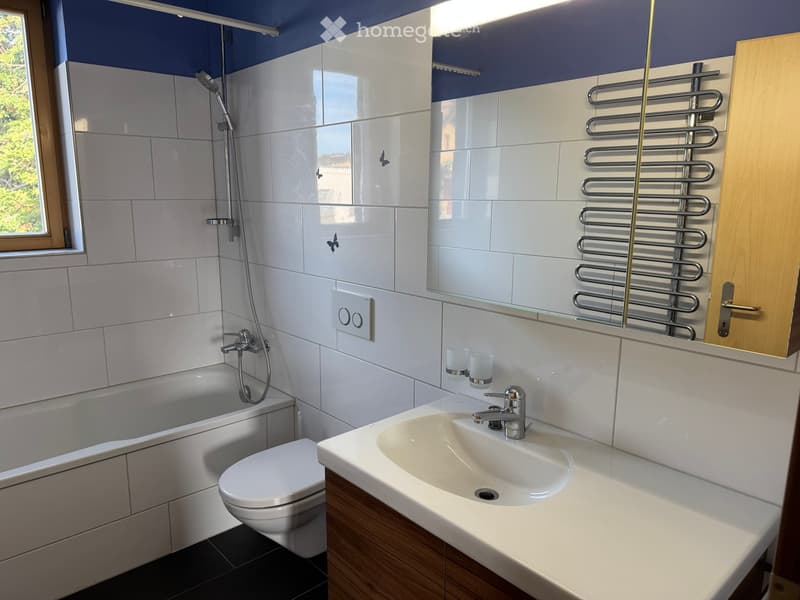 Modern bathroom with white tiles, a bathtub, toilet, sink, and a large mirror. Features a radiator and a window with a view of greenery.