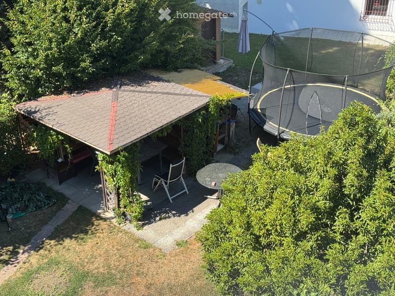 Garden area with a trampoline, patio furniture, covered seating area, and surrounding greenery.