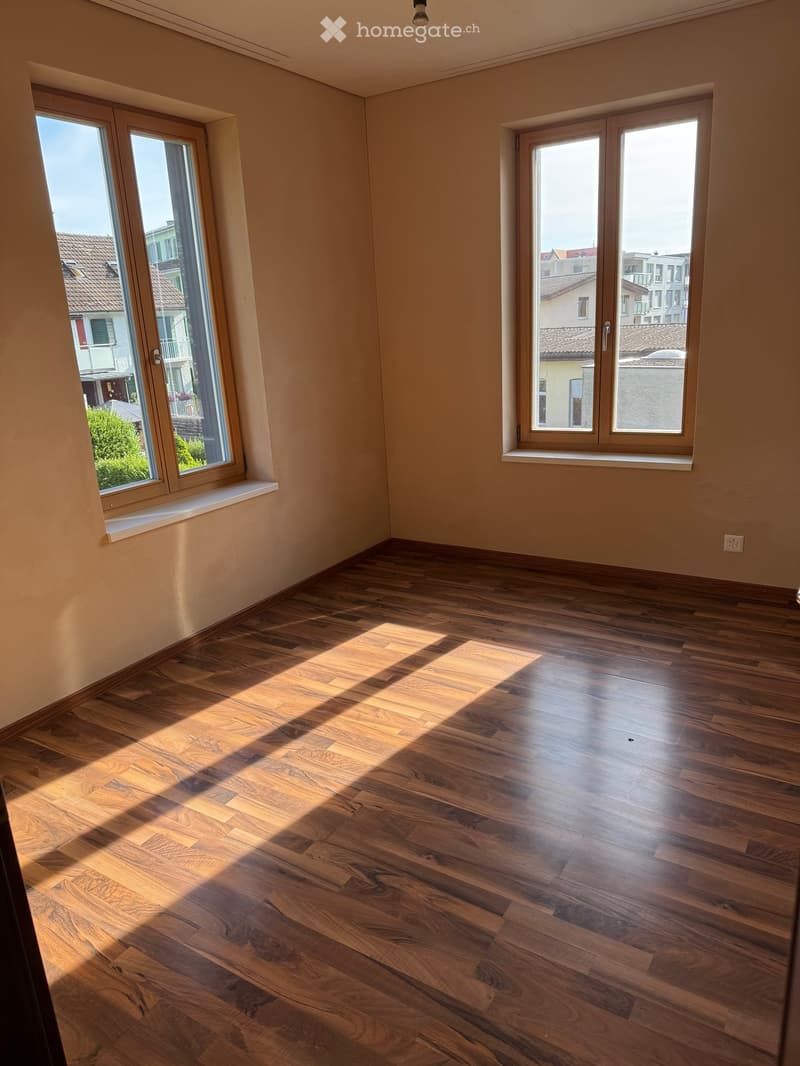 Empty room with wooden flooring and large windows letting in natural light. The walls are painted a light beige color, and the room has a clean, minimalist design.
