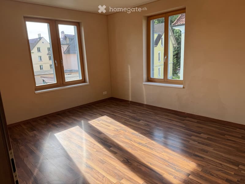 Empty room with wooden flooring and large windows, allowing natural light to fill the space. The walls are painted in a light beige color, and the room has a clean, minimalist design.