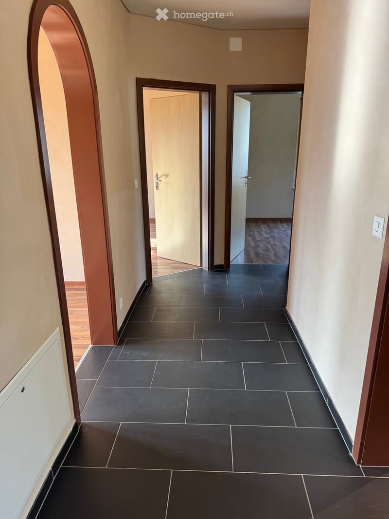 A clean, modern hallway with dark tiled flooring and light-colored walls. Features include an arched entryway, two open doors leading to adjacent rooms, and visible light switches and outlets.