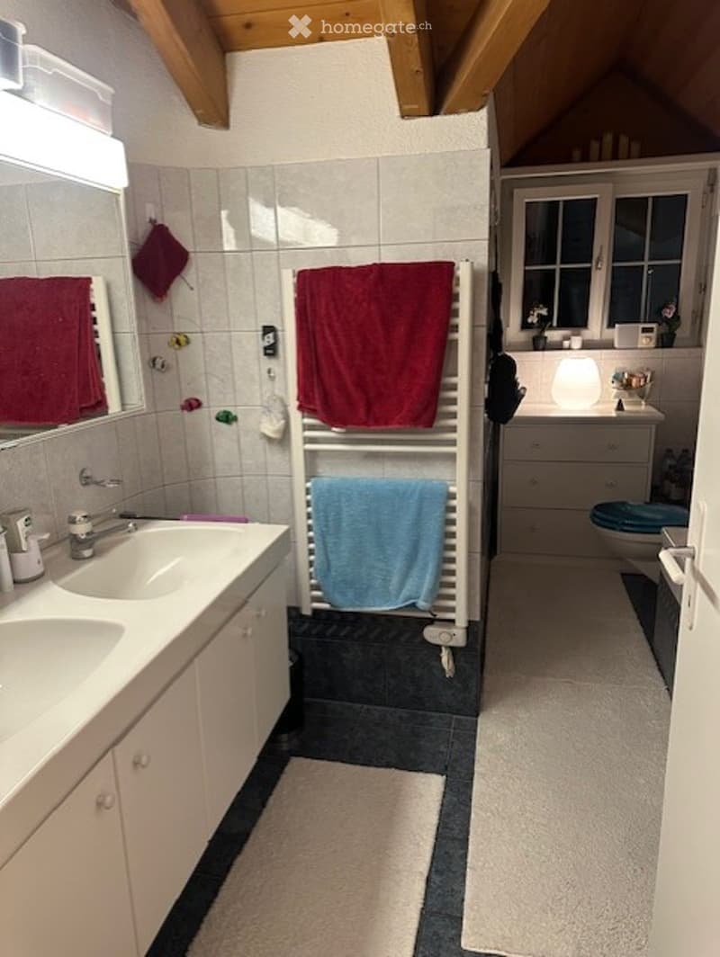 Bright bathroom with double sink, large mirror, towel rack with red and blue towels, white cabinets, and a toilet. The room features a slanted wooden ceiling and a window with white frames.
