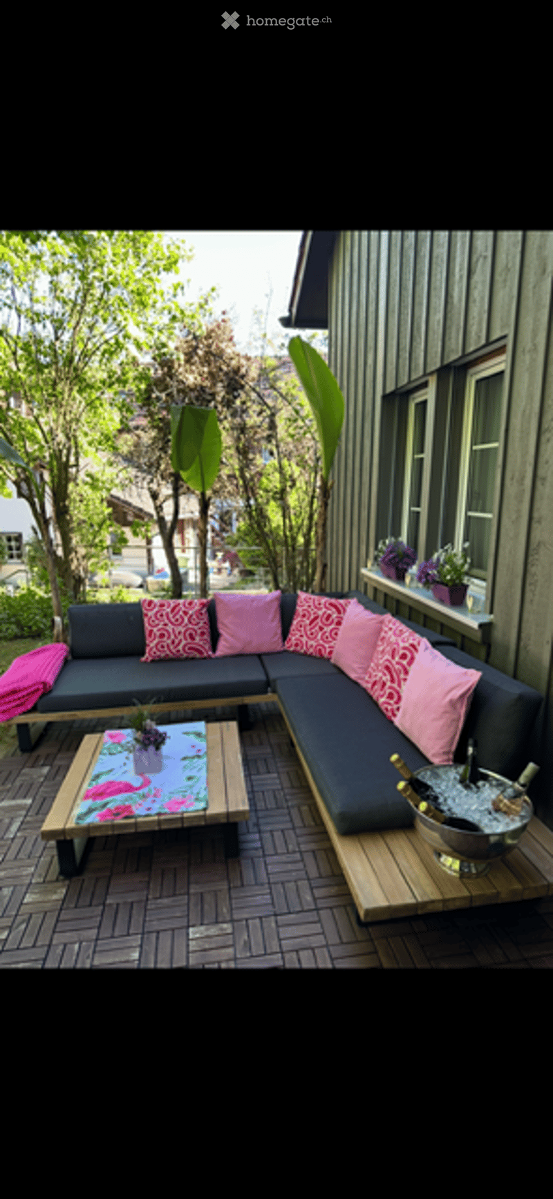 A modern outdoor seating area with a dark grey sectional sofa, pink and red patterned cushions, a wooden coffee table with a floral centerpiece, and a large ice bucket with champagne bottles. The area is surrounded by lush greenery and features a wooden d