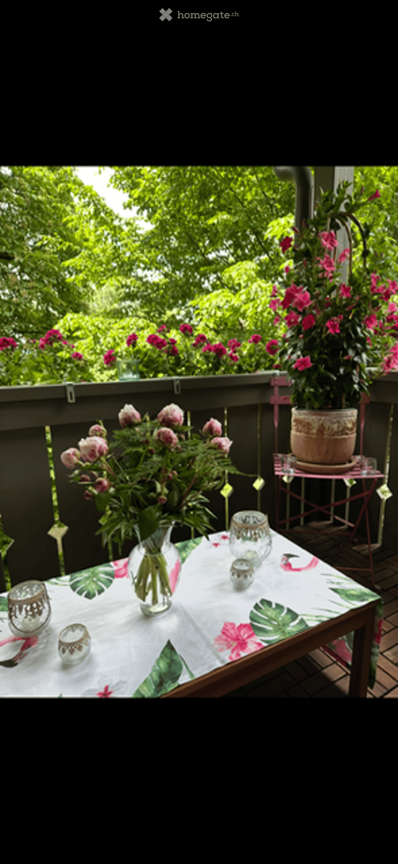 Balcony with a table covered in a floral tablecloth, surrounded by potted plants and flowers, with a railing and greenery in the background.