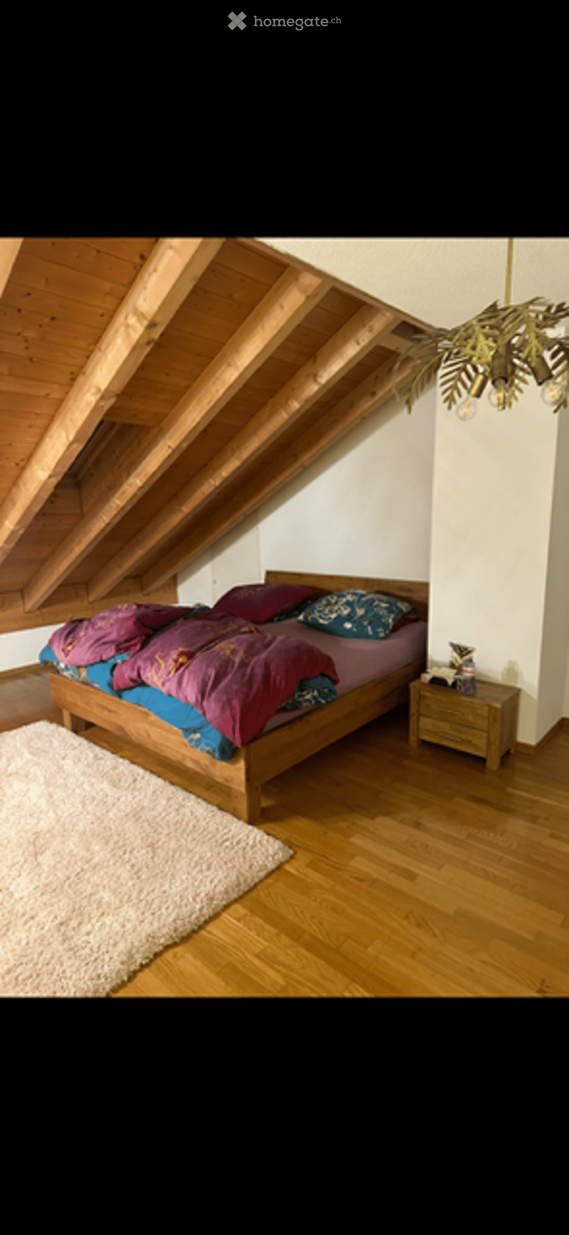 Cozy attic bedroom with wooden slanted ceilings, a large bed with colorful bedding, a nightstand, a fluffy white rug, and a hanging chandelier with a leafy design.