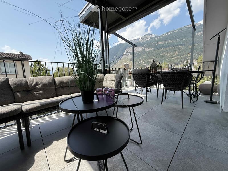 Balcony with a view of mountains, featuring a sofa, tables, chairs, and a potted plant.