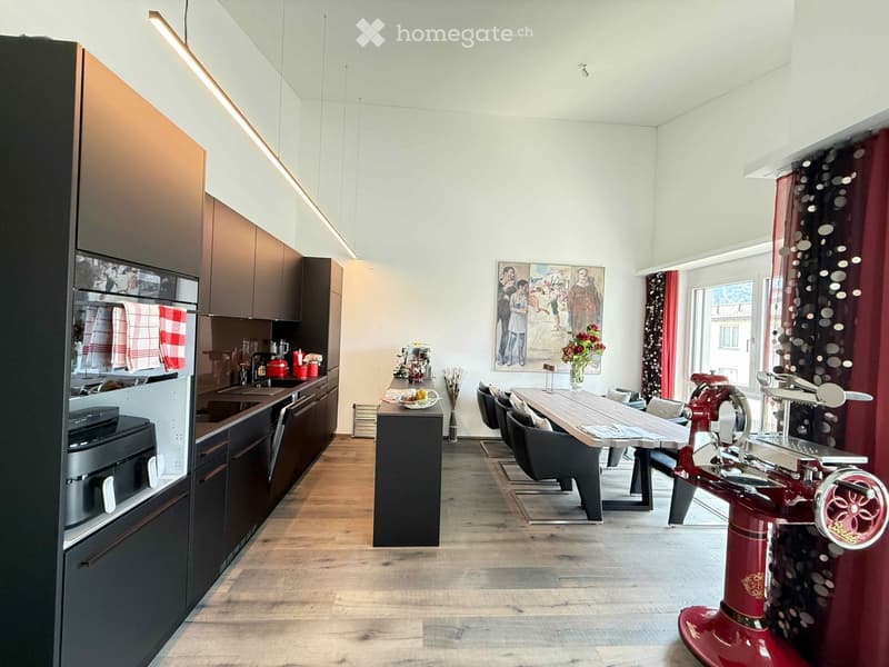 Modern kitchen with dark wooden cabinets, stainless steel appliances, a central island, and a dining area with a wooden table and chairs. Features a red sewing machine as a decorative element, a vase with flowers, and a painting on the wall.